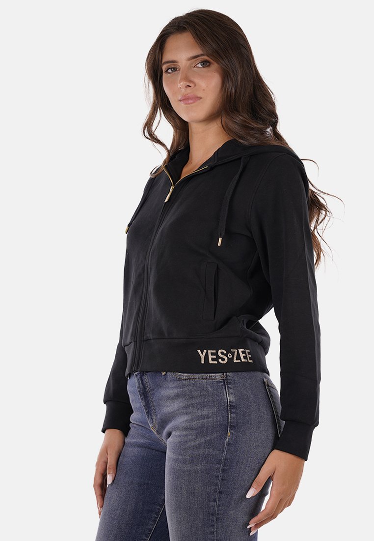 Felpa nera con zip realizzata in tessuto morbido, caratterizzata da un cappuccio, tasche frontali, dettagli della zip in oro e il logo "YES:ZEE" in un colore a contrasto.