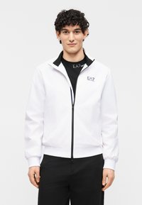 Νεαρός άνδρας με κοντά κατσαρά μαλλιά που φοράει λευκό μπουφάν EA7 με φερμουάρ πάνω από μαύρο μπλουζάκι EA7, στέκεται μπροστά σε ένα απλό λευκό φόντο.