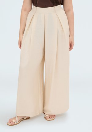 Vrouw draagt beige broek met hoge taille en wijde pijpen en lichtbeige platte sandalen, staand op een effen lichte achtergrond.