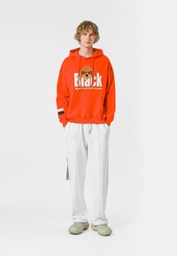 Sudadera roja con diseño gráfico que presenta un oso y texto en negrita, pantalones blancos y zapatos verdes texturizados. Estilo casual de streetwear.