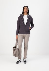 Sudadera con capucha y cremallera en gris oscuro con bolsillos frontales, llevada con una camiseta blanca, pantalones beiges y zapatillas negras, sosteniendo una bolsa gris.