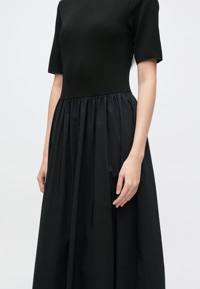 Čierne šaty s priliehavým živôtikom, krátkymi rukávmi a nariasenou, vzdušnou sukňou z textúrovaného materiálu. Celé s hladkým povrchom.