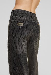 Fekete farmernadrág perforált mintával. Csillogó szegecs díszítésekkel és Lois márkajelzés a hátsó zsebén. Testhezálló fazon.