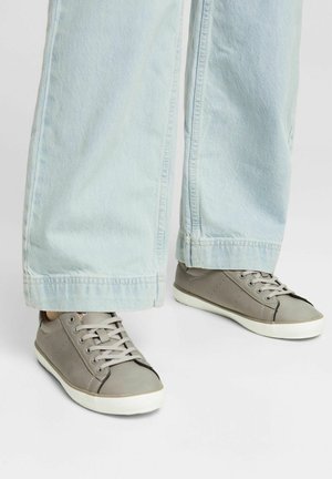 Person iført lyseblå vidde ben jeans og grå snøresko i afslappet stil, der står på en hvid overflade.