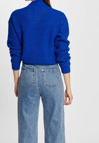Niebieski sweter z dzianiny o wysokim dekolcie i ściągaczami, w zestawieniu z jasnoniebieskimi, szerokimi dżinsami z wysokim stanem, posiadającymi tylne kieszenie.