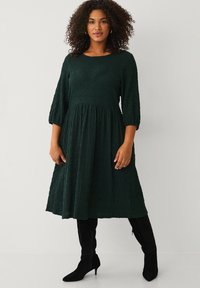 Mørkegrø dress med tekstur, rund halsudskæring, trekvartærmer og flagrende nederdel der når midt på læggen, sammen med knæhøje sorte støvler.