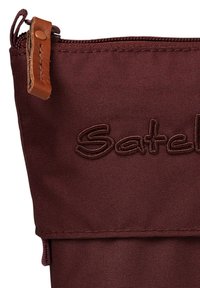 Pochette en tissu bordeaux avec logo "Satchel" en relief. Dotée d'une tirette en cuir marron et de coutures détaillées. Texture lisse.