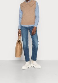 Gilet lavorato a maglia marrone con colletto ondulato sopra una camicia a righe blu e bianche, abbinato a jeans blu e stivaletti bianchi con suola spessa. Tiene una borsa marrone.