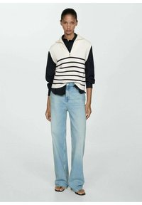 Gilet a maglia crema con righe blu navy, con colletto a zip, indossato sopra una camicia nera. Abbinato a jeans a gamba larga di un blu chiaro.