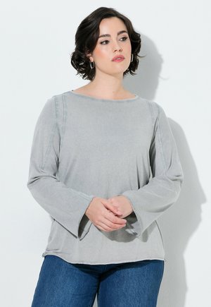Frau mit kurzen dunklen Haaren, die ein hellgraues Langarmshirt und blaue Jeans trägt, steht mit verschränkten Händen vor einer weißen Wand.
