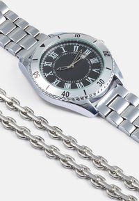 Ceas de mână de argint cu cadran cu numere romane, față neagră, fereastră pentru dată și brățară metalică cu link-uri, alături de o curea de argint.