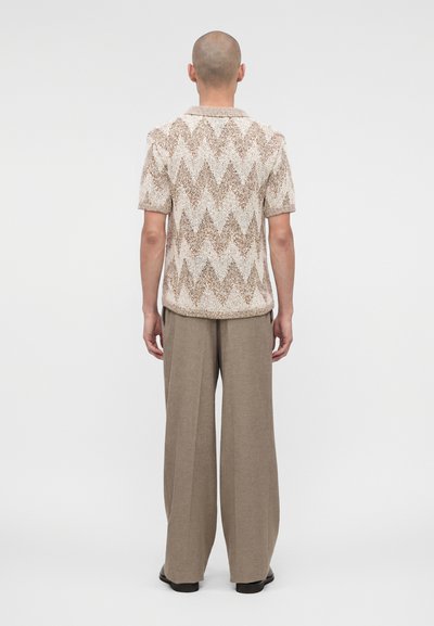Άνδρας με ξυρισμένο κεφάλι που φοράει μπεζ πλεκτό κοντομάνικο πουκάμισο με ζικ-ζακ σχέδιο και φαρδιά παντελόνια ταουπ, στέκεται με την πλάτη του προς τα εμπρός.