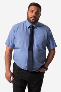 Mand med kort mørkt hår og skæg iført en lyseblå kortærmet skjorte, mørkt slips, sorte bukser og et armbåndsur, står med den ene hånd i lommen.