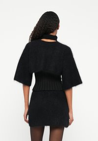 Μαύρο, φουντωτό πλεκτό πουλόβερ με ψηλό ριμπ γιακά, κοντά μανίκια και εφαρμοστή υφασμένη ζώνη, φορεμένο πάνω από μίνι φούστα του ίδιου χρώματος.