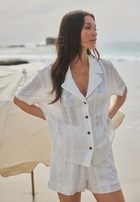 Chemise boutonnée légère en lin blanc et shorts assortis avec un motif texturé. Comprend un col, des poches latérales et des boutons marron.