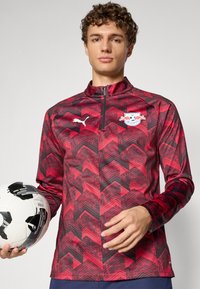 Rød og svart mønstret treningsjakke med halv-glidelås, med PUMA-logo og lagmerke. Laget av et glatt, lett materiale.