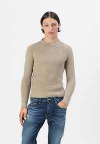 Maglione lavorato a maglia beige con scollatura rotonda e maniche lunghe, abbinato a jeans blu con un leggero effetto strappato. Texture semplice.