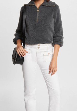 Femme portant un pull gris foncé en tissu duveteux avec une fermeture éclair dorée, un pantalon blanc taille haute avec des boutons, et portant un sac bandoulière noir.