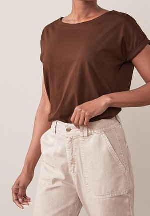 Personne portant une chemise marron à manches courtes rentrée dans un pantalon taille haute beige clair avec un bouton et une taille élastique.