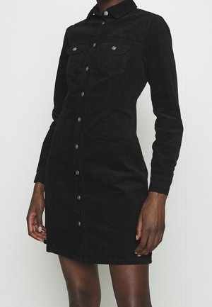 Kvinde iført en sort corduroy skjortekjole med knapper, lange ærmer og brystlommer, stående mod en ensfarvet lys baggrund.