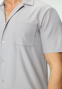 Chemise à manches courtes gris clair avec col et poche plaquée. Le tissu a une texture légèrement côtelée et présente des boutons sur le devant.