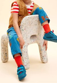 Las manos de un niño reposan sobre una silla blanca moteada y texturizada. Lleva un chaleco marrón, una camisa a rayas, pantalones azules con patrones coloridos y zapatos azules.