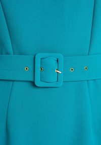 Podrobnost tyrkysových šatů s páskem, který má široký látkový pásek s obdélníkovou sponou a hladkou texturu látky v jednobarevném provedení.