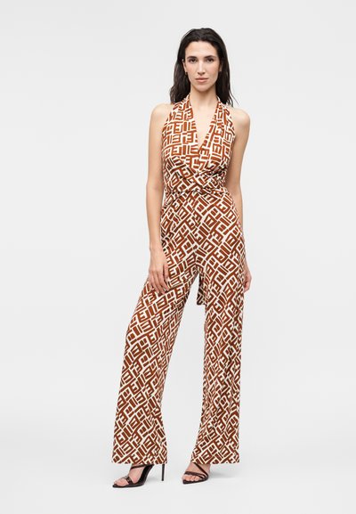 Γυναίκα που φοράει αμάνικη ολόσωμη φόρμα με halter neck και καφέ και λευκά γεωμετρικά σχέδια, φορώντας μαύρα σανδάλια με ψηλό τακούνι.