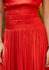 Robe plissée rouge avec une taille froncée. Confectionnée en tissu lisse et brillant, présentant des plis verticaux et une couleur vibrante et uniforme.