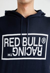 Marineblå hettegenser med hvit strikket tekst som sier "RED BULL RACING" i en ramme, med snøring i hetten og myk tekstur.
