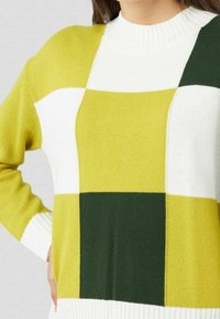 Pull en maille à col rond, avec un motif en damier vert, jaune et blanc. Poignets côtelés. Texture douce, épaisseur moyenne.