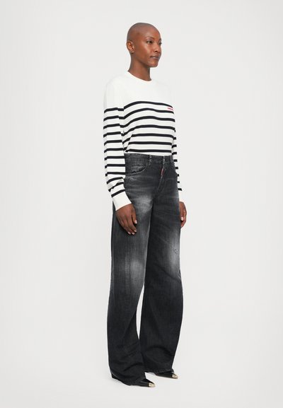 Woman with shaved head wearing a white and black striped long-sleeve shirt, faded wide-leg black jeans, and black pointed toe shoes.