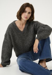 Grijze V-hals trui met losse, textuur gebreide stof en oversized mouwen, gecombineerd met high-waisted blauwe jeans en zwarte sandalen met banden.