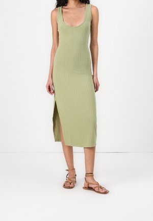 Kvinde iført en ærmeløs, ribbet lys grøn midi-kjole med sideslids og brune flade sandaler med stropper.