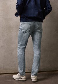 Persoon draagt lichtblauwe spijkerbroek, witte sneakers en een marineblauwe jas, staat met handen in de zakken tegen een kale betonnen muur.