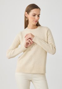 Suéter de punto beige con puños y dobladillo acanalados, cuello redondo y textura suave, combinado con pantalones de color claro, con las manos unidas al frente.