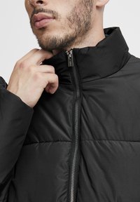 Black padded jacket with a high collar, featuring a front zipper and subtle ribbed texture. The fabric appears smooth and lightweight.
