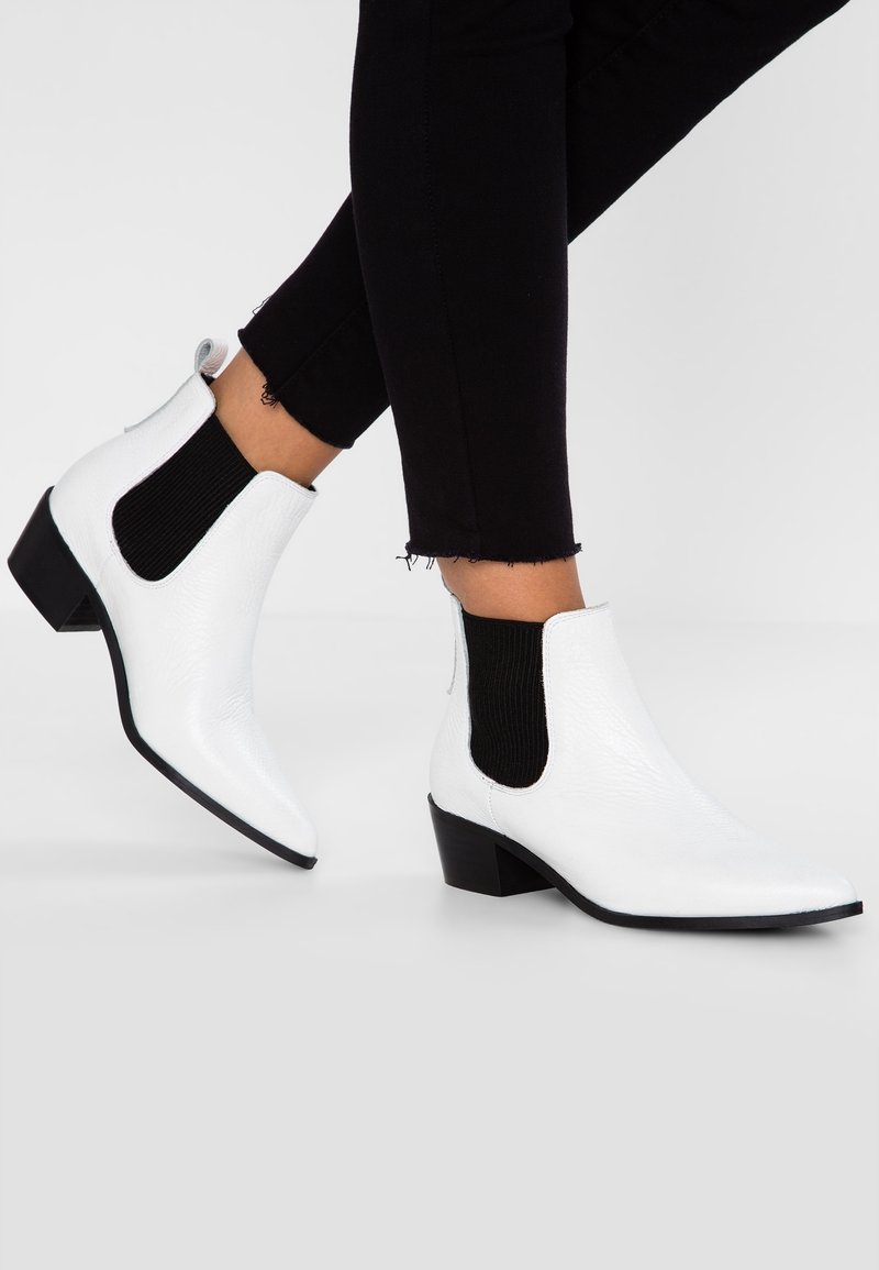 Bottes blanches à bouts pointus et chevilles basses avec panneaux élastiques noirs sur les côtés, portées avec un pantalon noir court.