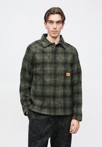 Młody mężczyzna z kolczykami, ubrany w zielono-czarną kraciastą zapinaną na guziki kurtkę i ciemne spodnie z wzorem, stojący z jedną ręką w kieszeni.