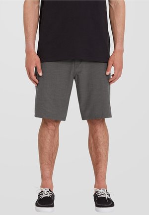 Homme portant un short gris foncé jusqu'aux genoux, une chemise noire et des baskets noires avec des lacets blancs, debout devant un fond uni.