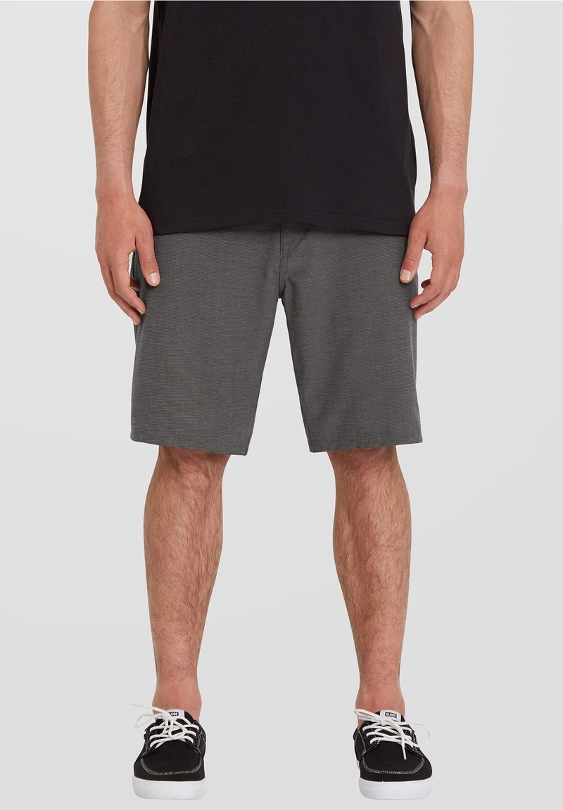 Homme portant un short gris foncé jusqu'aux genoux, une chemise noire et des baskets noires avec des lacets blancs, debout devant un fond uni.