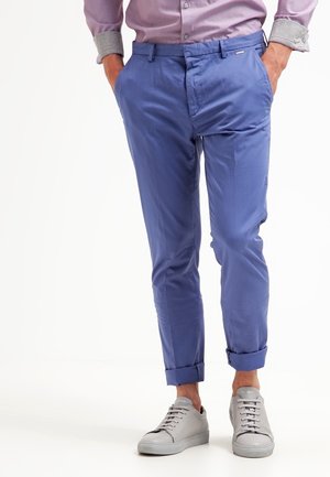 Homme portant un chino bleu retroussé, des baskets gris clair et une chemise rose aux manches retroussées, les mains dans les poches.