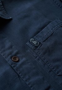 Marineblauw overhemd met knopen en een borstzak, voorzien van een bruine knop en een ronde logopatch met de tekst "Super Dry". Gestructureerde stof.