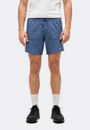Mann trägt blaue Nike Sportshorts mit Kordelzug, weiße Socken und schwarze Laufschuhe, steht mit entspannten Armen an den Seiten.