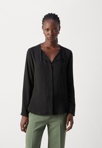 Blusa nera a maniche lunghe con scollatura a V e chiusura con bottoni, realizzata in tessuto morbido. Indossata con pantaloni verdi, dotati di una vestibilità comoda.