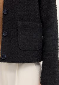 Schwarze, strukturierte Jacke mit cropped Design, ausgestattet mit zwei blauen Knöpfen und einer einzelnen aufgesetzten Tasche auf der Vorderseite. Weißer Stoff ist darunter sichtbar.