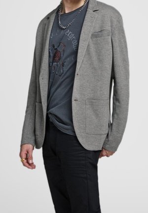 Homme portant un blazer gris par-dessus un t-shirt graphique sombre, un pantalon noir, un collier chaîne et des bagues aux doigts, sur un fond uni.