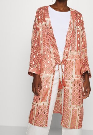 Osoba nosząca różowy kimono w wzory ze złotymi akcentami, zawiązane z przodu frędzlami, nałożone na biały top i białe spodnie.