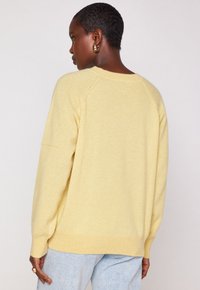 Donna con i capelli corti che indossa orecchini a cerchio d'oro, un maglione giallo chiaro e jeans blu chiaro, rivolte di spalle su uno sfondo semplice.