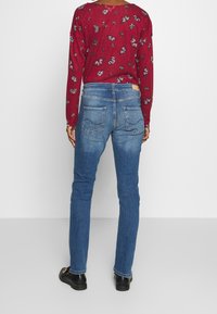 Red long-sleeve top with butterfly print, paired with fitted blue jeans featuring back pockets and a slightly distressed hem. Black shoes.